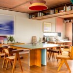 Štýlová jedáleň s veľkým štvorcovým stolom, ikonickými drevenými stoličkami s výpletom a červeným závesným svietidlom.