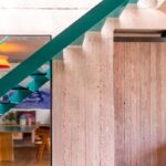 Tyrkysové kovové schodisko vedľa ružovej betónovej steny s textúrou dreveného debnenia a zrkadlovou plochou.