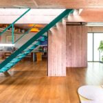 Interiér s výrazným tyrkysovým schodiskom, zrkadlovou stenou, drevenou podlahou a ružovým betónovým pilierom.
