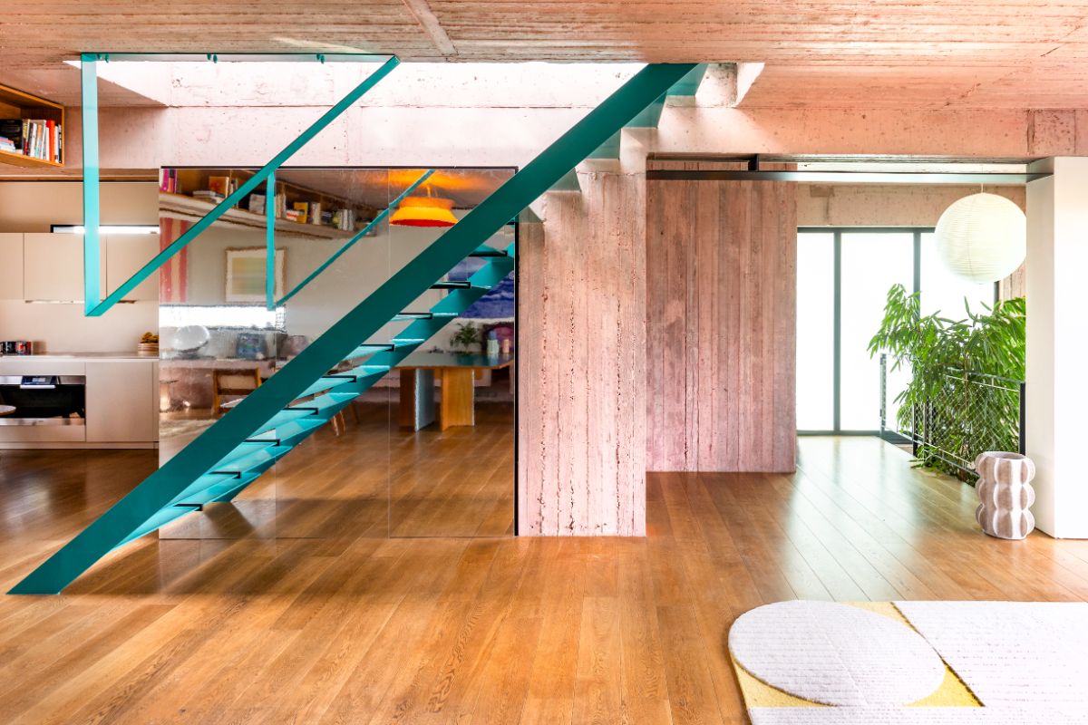 Interiér s výrazným tyrkysovým schodiskom, zrkadlovou stenou, drevenou podlahou a ružovým betónovým pilierom.