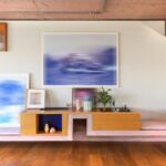 Nástenná kompozícia s modrými abstraktnými obrazmi, drevenou policou a ružovou betónovou lavicou s mäkkým čalúnením.