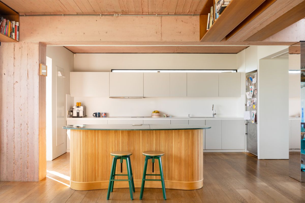 Minimalistická biela kuchynská linka s dreveným lamelovým ostrovom a zelenými barovými stoličkami v modernom byte.