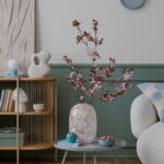 Sivý okrúhly stolík s kvitnúcimi čerešňovými vetvičkami vo váze, zeleným stenovým panelom a bielym oblým kreslom.