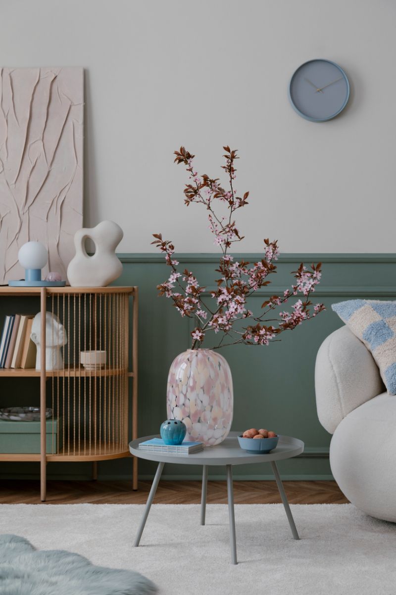 Sivý okrúhly stolík s kvitnúcimi čerešňovými vetvičkami vo váze, zeleným stenovým panelom a bielym oblým kreslom.