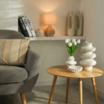 Drevený príručný stolík s bielymi tulipánmi vo váze, pletenou stolnou lampou a sivým čalúneným kreslom v rohu.