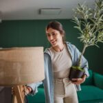 Žena v svetlomodrej košeli drží zelený olivovník v čiernom kvetináči pri drevenej lampe.