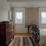 Spálňa s kovovou posteľou, kamenným obkladom steny a starožitnou drevenou komodou pri okne.