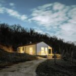 Biely jednopodlažný dom so sedlovou strechou a veľkými presklenými oknami v kopcovitom teréne.