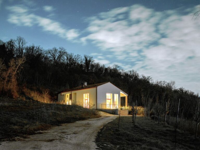 Biely jednopodlažný dom so sedlovou strechou a veľkými presklenými oknami v kopcovitom teréne.