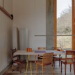 Masívny drevený stôl s rôznymi retro stoličkami pri vysokom okne.