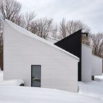 Moderný biely dom s ostrou šikmou strechou a čiernym akcentom v zasneženej lesnej krajine.