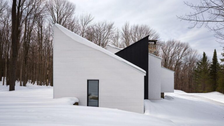Moderný biely dom s ostrou šikmou strechou a čiernym akcentom v zasneženej lesnej krajine.