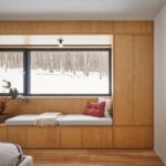 Drevený okenný výklenok s mäkkým sedením, zelenou rastlinou a výhľadom na zasnežený les.