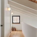 Podkrovná chodba s bielymi šikmými trámami, drevenou podlahou a guľatými nástennými svietidlami.