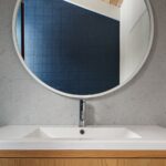 Biele keramické umývadlo na drevenej skrinke s kruhovým zrkadlom a sivým kamenným obkladom.