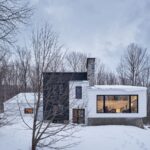 Moderný dvojpodlažný dom s čiernym a bielym obkladom v zasneženej krajine s vysokými stromami.