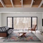 Obývacia izba s čiernou koženou sedačkou, farebným kobercom a kamenným kozubom pod dreveným stropom.