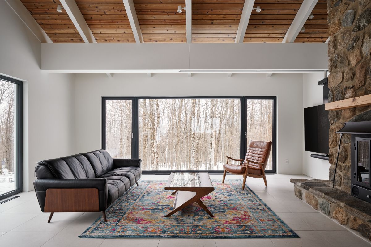 Obývacia izba s čiernou koženou sedačkou, farebným kobercom a kamenným kozubom pod dreveným stropom.