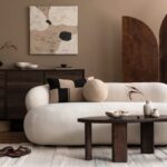 Biela zaoblená buclé pohovka, tmavý drevený konferenčný stolík a abstraktný béžový obraz na hnedej stene.