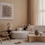 Krémová modulárna pohovka, nízky drevený stolík, prútené kreslo a plastický textilný obraz v pieskovom interiéri.