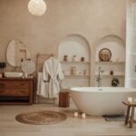 Voľne stojaca biela vaňa, oblúkové niky s policami, drevená komoda a okrúhle jutové zrkadlo v zemitej kúpeľni.