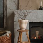 Béžová organická váza na drevenom stolčeku pred sivým betónovým krbom s pleteným košom na drevenej podlahe.