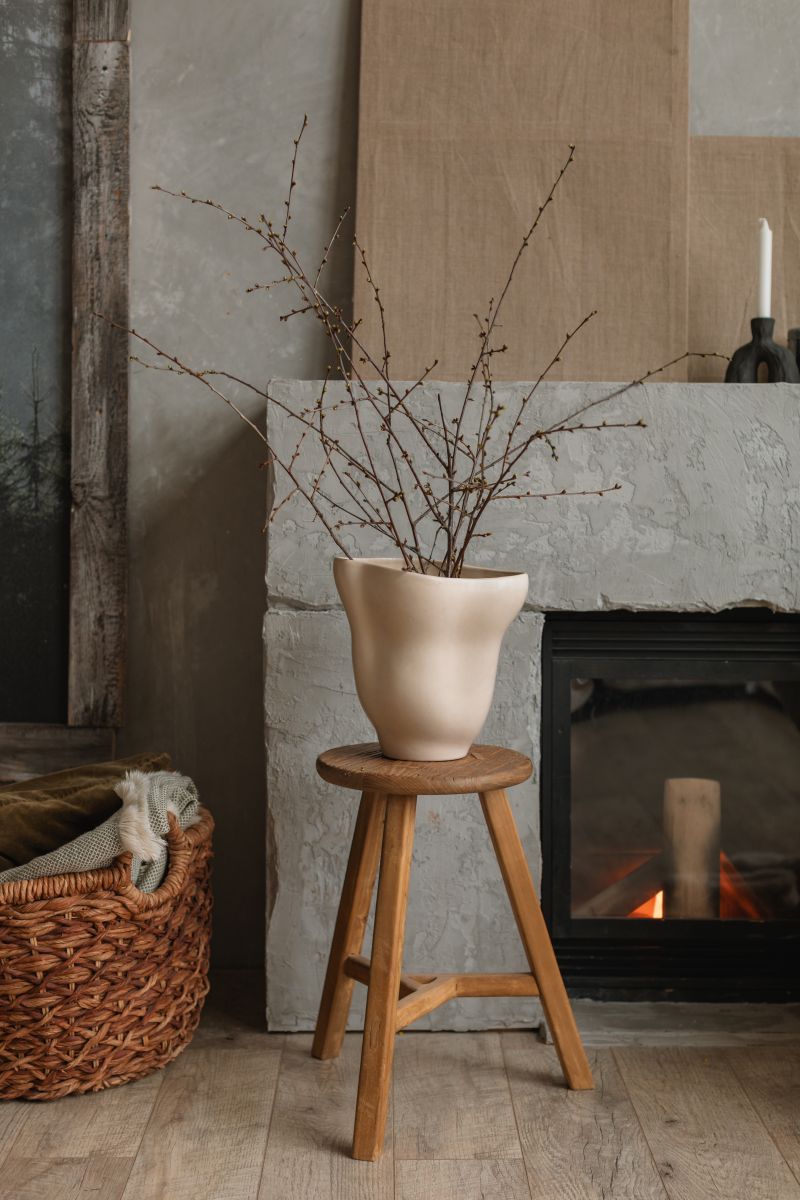 Béžová organická váza na drevenom stolčeku pred sivým betónovým krbom s pleteným košom na drevenej podlahe.