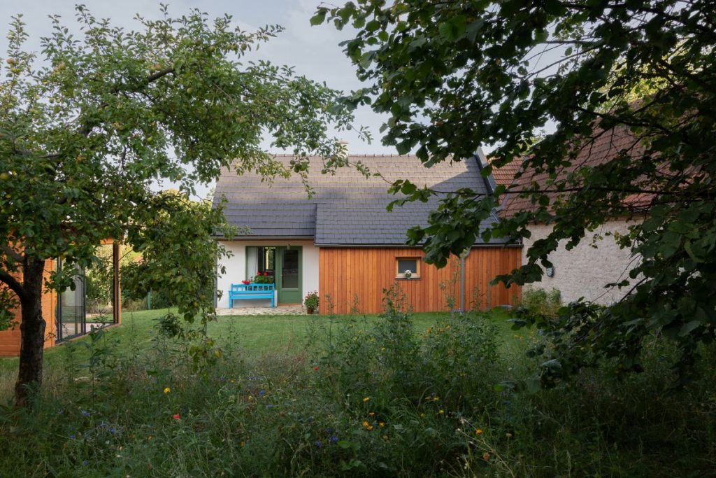 Záhrada so zrekonštruovanou chalupou s drevenou a bielou fasádou a zelenými oknami na vidieku.