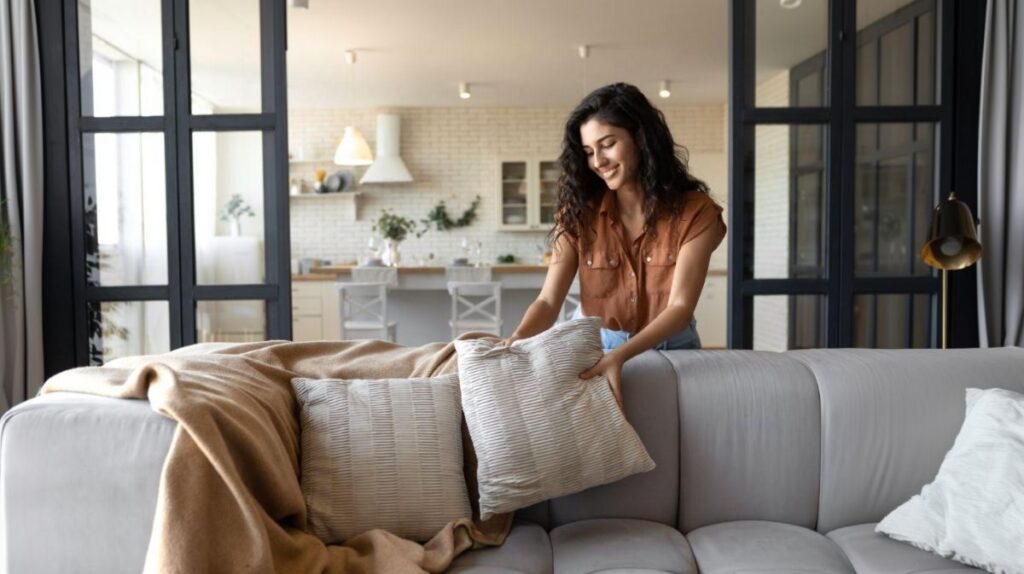 Žena upravuje vankúše na sivej pohovke s béžovou vlnenou dekou a v modernom interiéri.
