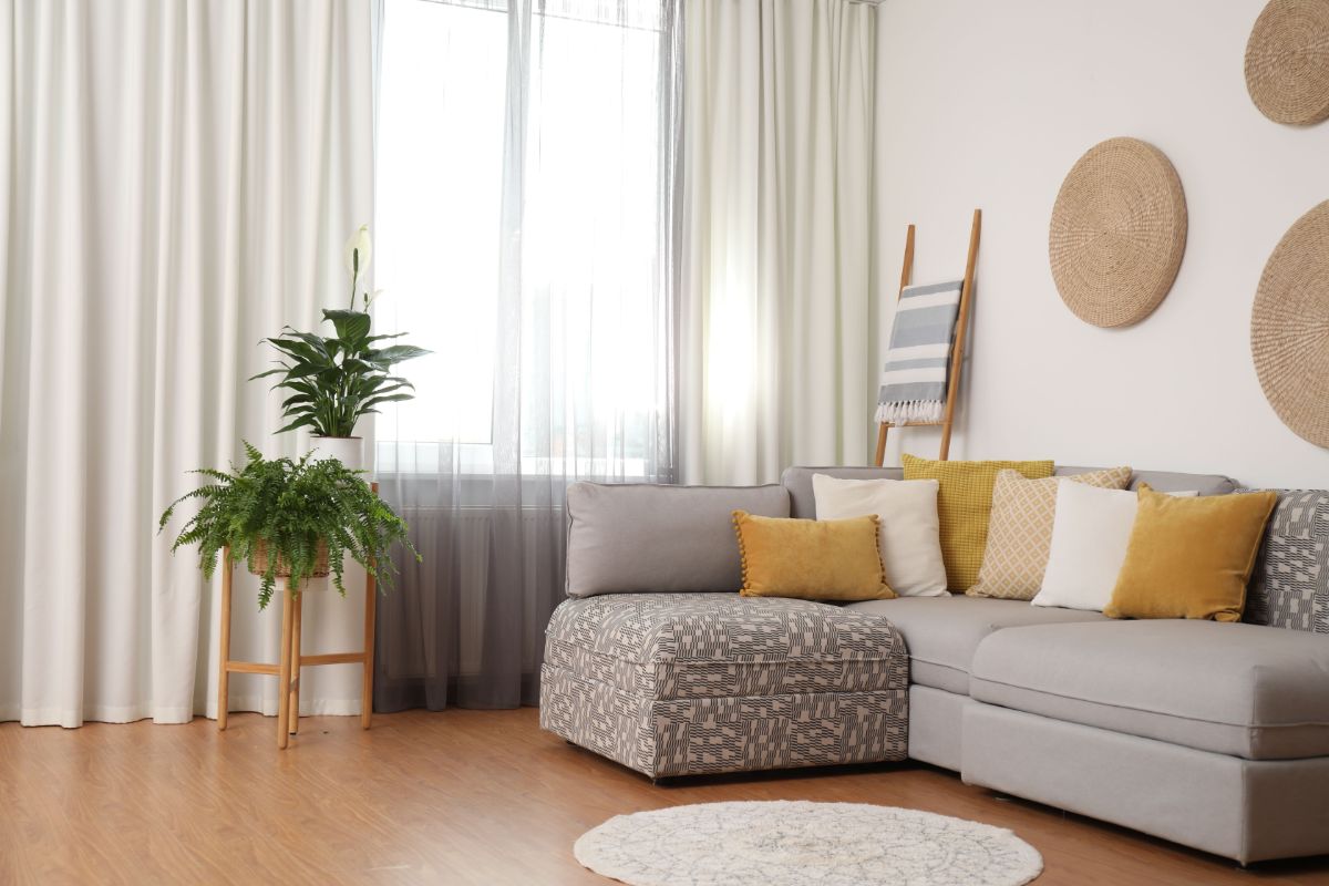 Rohová sivá pohovka so žltými dekoratívnymi vankúšmi pri okne s priesvitnými záclonami a závesmi.