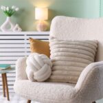 Biele bouclé kreslo so smotanovým vankúšom v tvare uzla a ďalším smotanovým vankúšom s rebrovaným vzorom.