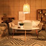 Béžová zaoblená sedačka s geometrickým vzorovaným kobercom a závesným prúteným svietidlom, vydávajúcim teplé žlté svetlo.