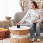 Mladá žena sediaca na sivej sedačke nastavuje bluetooth reproduktor, položený na okrúhlom ratanovom stolíku.