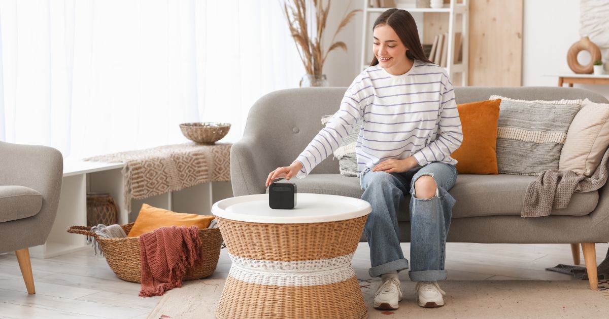 Mladá žena sediaca na sivej sedačke nastavuje bluetooth reproduktor, položený na okrúhlom ratanovom stolíku.