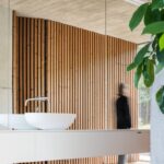 Moderné biele umývadlo pred zrkadlovou stenou s odrazom dreveného lamelového obkladu.