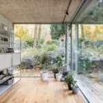 Kovový policový regál s knihami pri veľkoformátovom presklení s výhľadom na kamennú skalku a les.