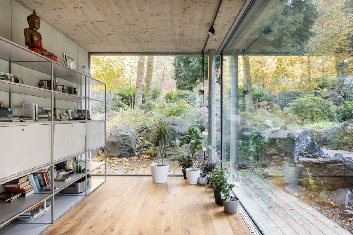 Kovový policový regál s knihami pri veľkoformátovom presklení s výhľadom na kamennú skalku a les.