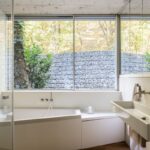 Biela vstavaná vaňa pod oknom s výhľadom na gabiónovú stenu a jesenné lístie,