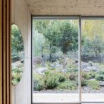 Veľké okno v betónovej stene s výhľadom na záhradu s ihličnatými stromami.