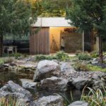 Moderný nízky dom s betónovým prekladom a veľkoformátovým presklením vedľa prírodného jazierka s tmavými kameňmi.