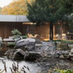 Pohľad cez potok s kamenným brehom na presklenú fasádu a minimalistický interiér s kamennou stenou.