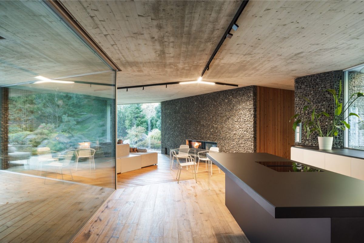 Interiér Banánky s čiernym kuchynským ostrovom, kozubom v kamennej stene a béžovou sedačkou pri presklenej fasáde.