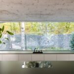 Široké pásové okno nad bielou kuchynskou linkou s výhľadom na gabiónový plot a jesennú zeleň.