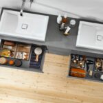 Sivá kúpeľňová skrinka s bielymi keramickými umývadlami a otvorenými zásuvkami s dreveným organizérom.