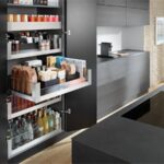 Vysoká čierna potravinová skriňa vnútornými vysúvacími skrinkami, plnými balených potravín a nápojov.