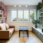 Hnedá čalúnená pohovka pred ružovou policovou priečkou, tmavozelená nízka skrinka a svetlá drevená podlaha pod oknom.