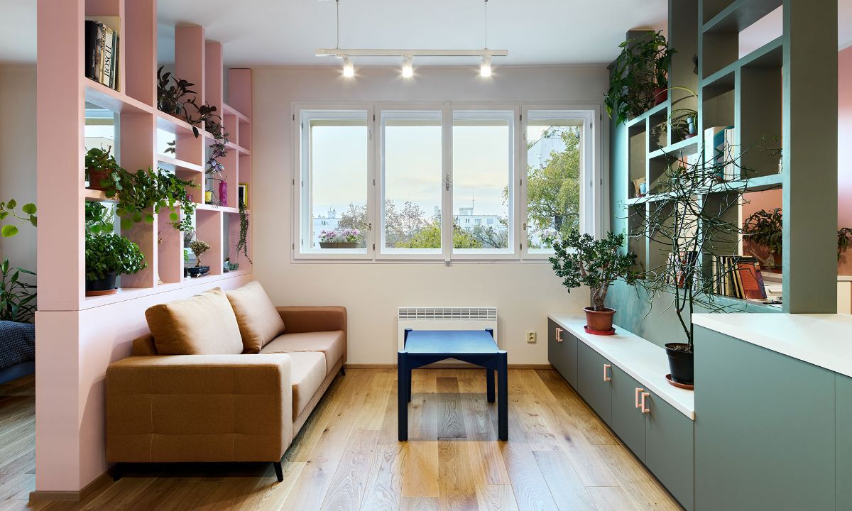 Hnedá čalúnená pohovka pred ružovou policovou priečkou, tmavozelená nízka skrinka a svetlá drevená podlaha pod oknom.