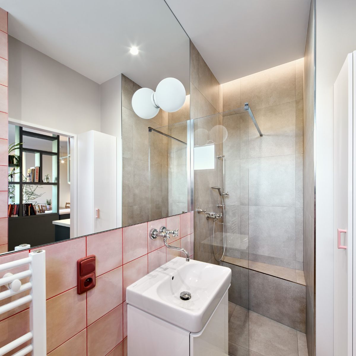 Kúpeľňa s bielym umývadlom na skrinke, ružovým obkladom veľkým zrkadlom a sprchovým kútom.
