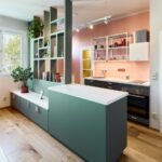 Tmavozelená kuchynská linka s ružovými úchytkami pod ružovou stenou, biela pracovná doska a otvorená kovová polica.