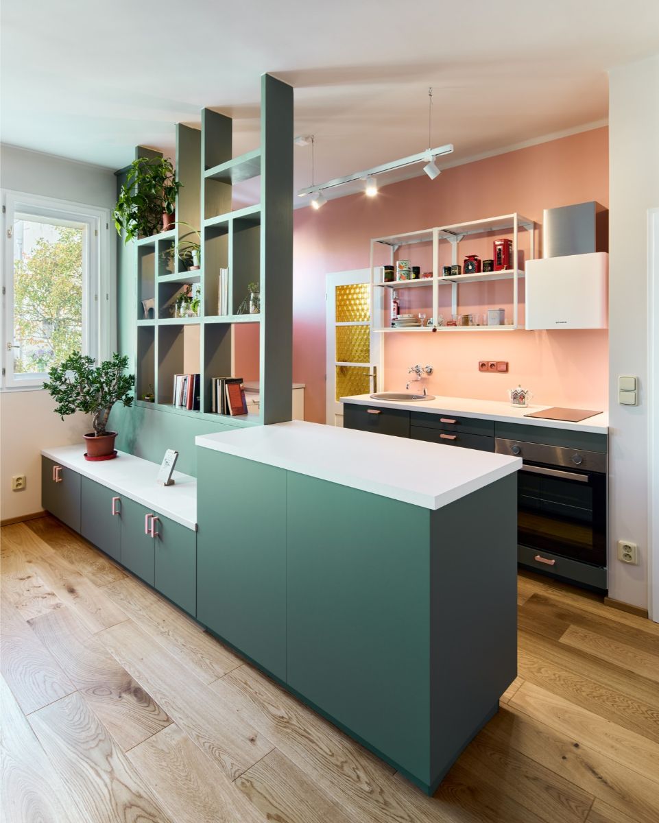 Tmavozelená kuchynská linka s ružovými úchytkami pod ružovou stenou, biela pracovná doska a otvorená kovová polica.